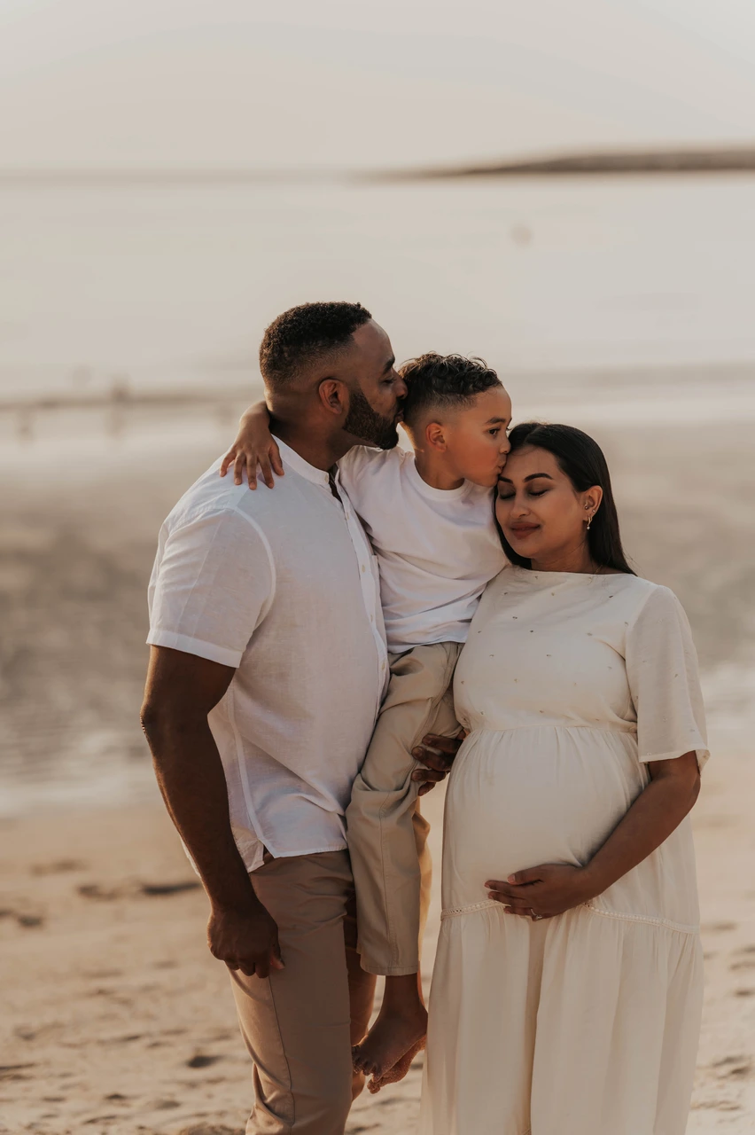 Maternity photoshoot with siblings - close family beach cuddle Parents and their child share a close beach cuddle around the baby bump in Dubai during a maternity photoshoot with siblings at golden hour.