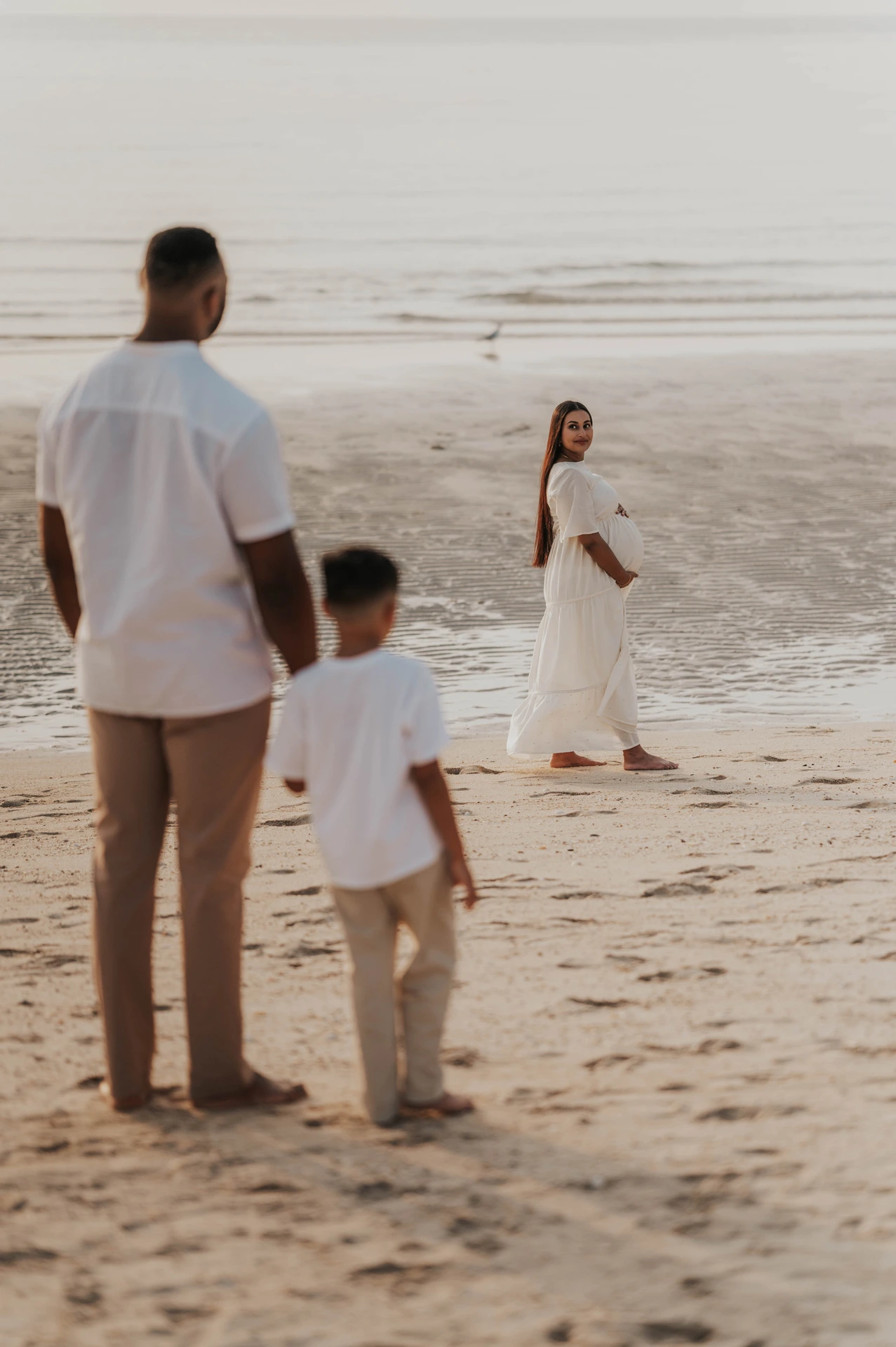 A toddler walks toward a pregnant mother at the waterline in Dubai while the family is photographed during a maternity photoshoot with siblings.