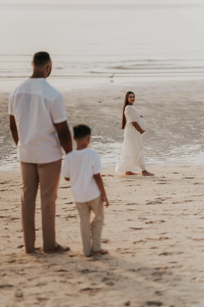 Maternity photoshoot with siblings - toddler beach approach frame A toddler walks toward a pregnant mother at the waterline in Dubai while the family is photographed during a maternity photoshoot with siblings.