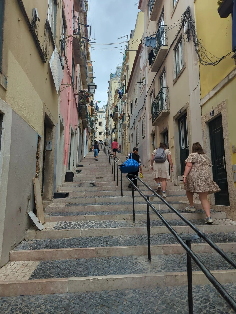 Lisbon Family Photoshoot - Steep Old-City Stairs Adults climb a long stone staircase between colourful Lisbon buildings, illustrating terrain to assess before a family photoshoot.