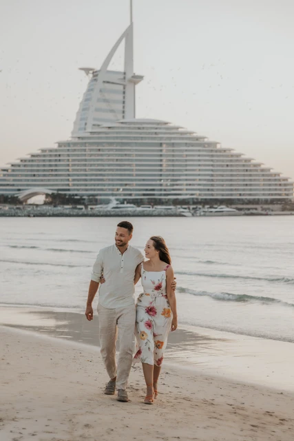A couple walks together on a Dubai beach with Jumeirah Beach Hotel visible behind them in soft morning haze.