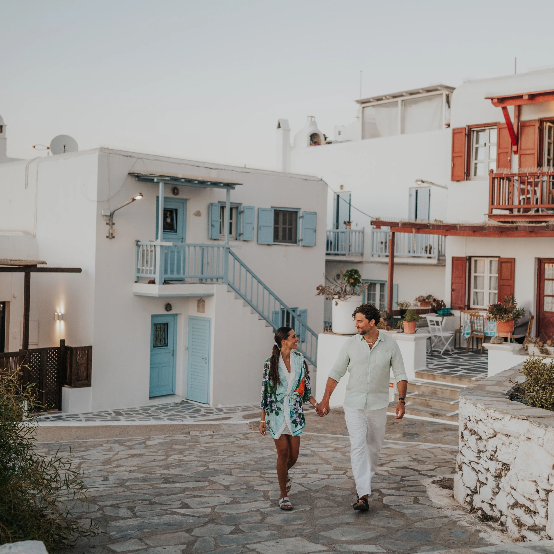 A Walking Approach Through Mykonos Town Two people walk hand in hand through Mykonos Town, surrounded by whitewashed houses with blue and red shutters.