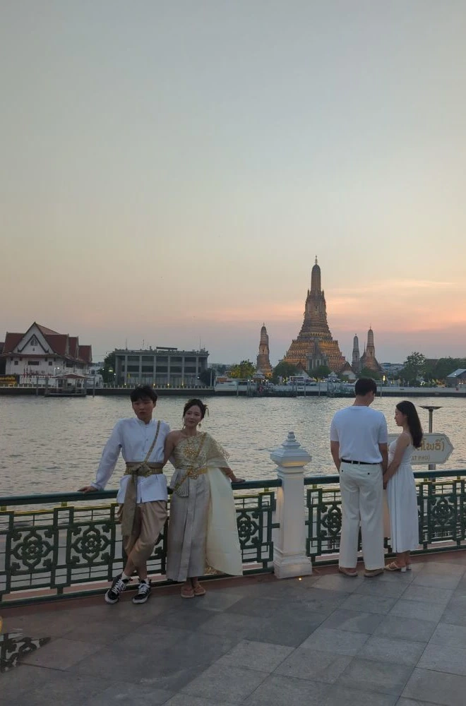 Bangkok Temple Outfit: Thai-Style Dress Two adults in white-and-gold Thai-style outfits stand beside Bangkok’s river with Wat Arun behind them, illustrating a Thai-style temple photoshoot look.