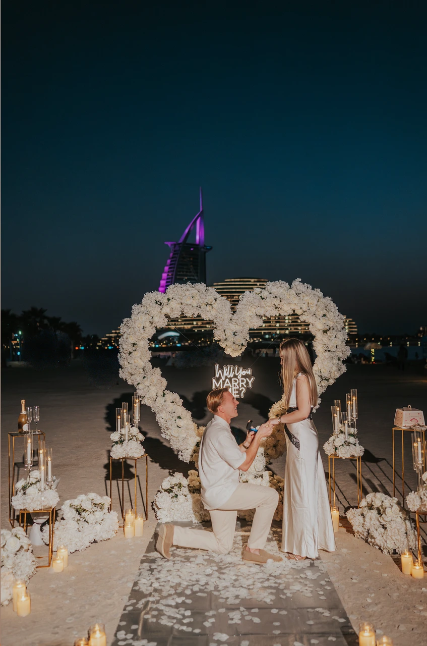 Heart Proposal Setup Dubai - white arch proposal at blue hour A man kneels to propose beneath a white floral heart setup on Dubai beach at blue hour, with Burj Al Arab visible beyond the water.