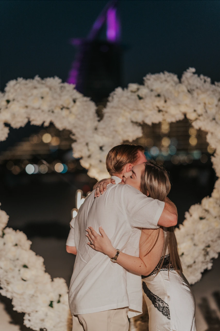 Heart Proposal Setup Dubai - couple hug in white heart setup A couple hugs after the proposal inside a white floral heart setup on Dubai beach at night, with warm lights and soft bokeh behind.