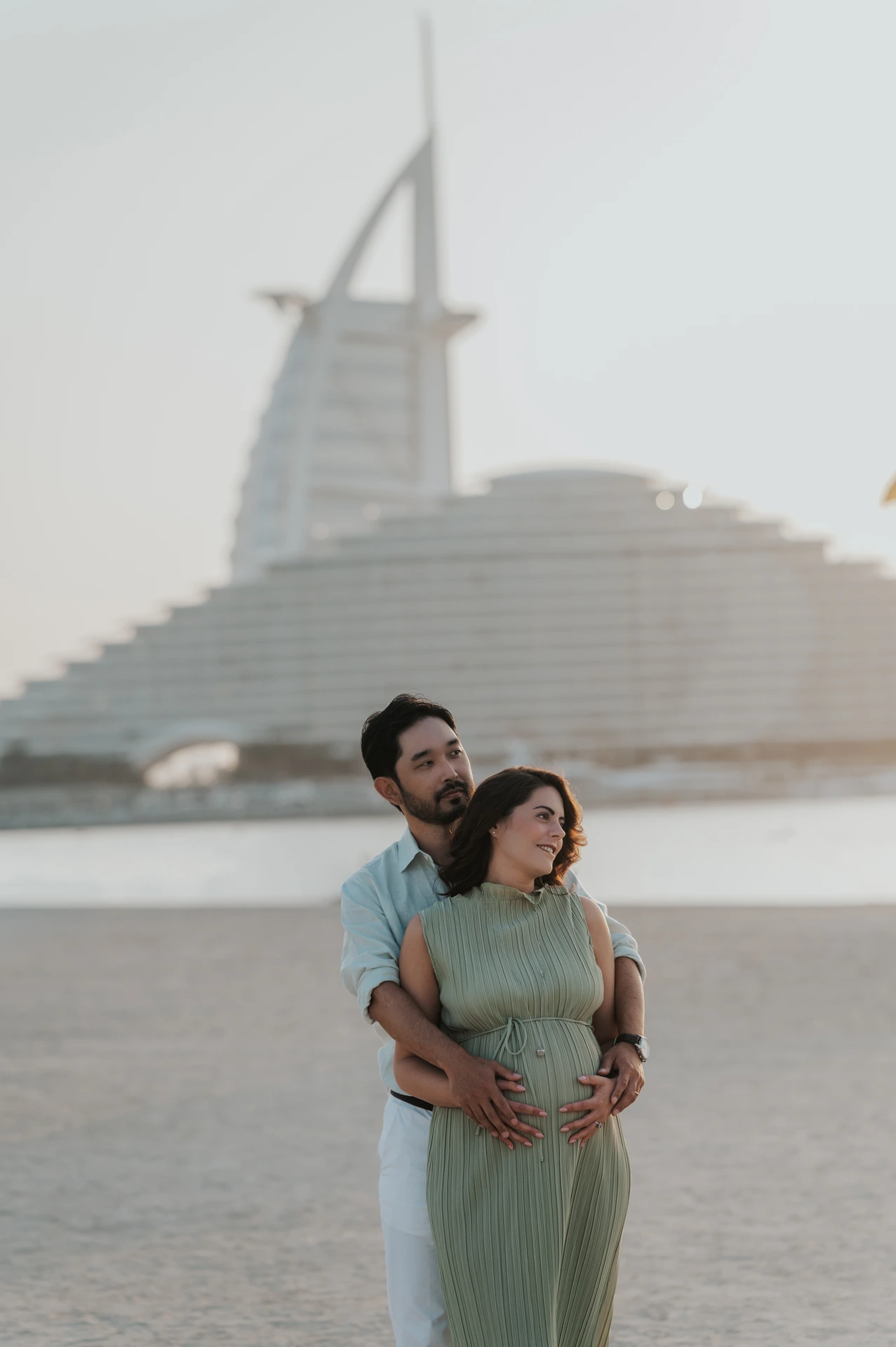 A man embraces his pregnant partner in a sage-green dress with Burj Al Arab behind them during a Dubai maternity photoshoot.