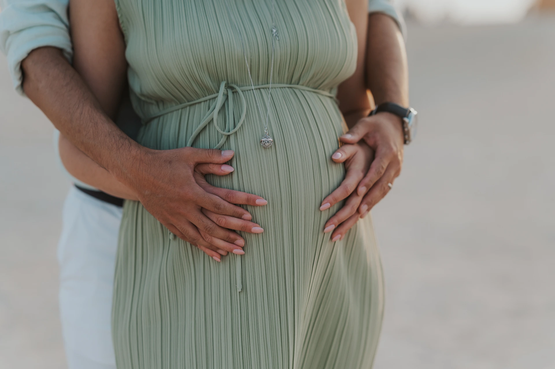 An expectant couple places both sets of hands around the visible bump beneath a sage-green maternity dress during a Dubai beach portrait.