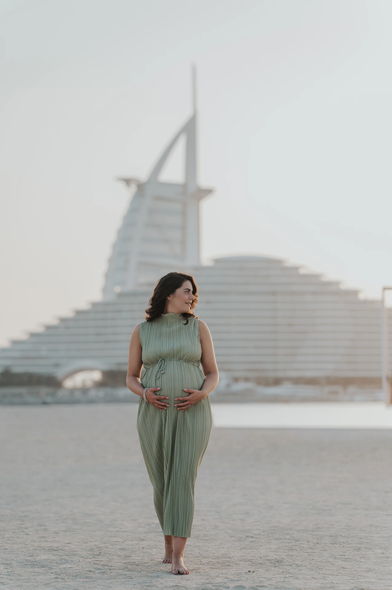 A pregnant woman in a sage-green dress holds her bump while posing with Burj Al Arab behind her during a Dubai maternity shoot.