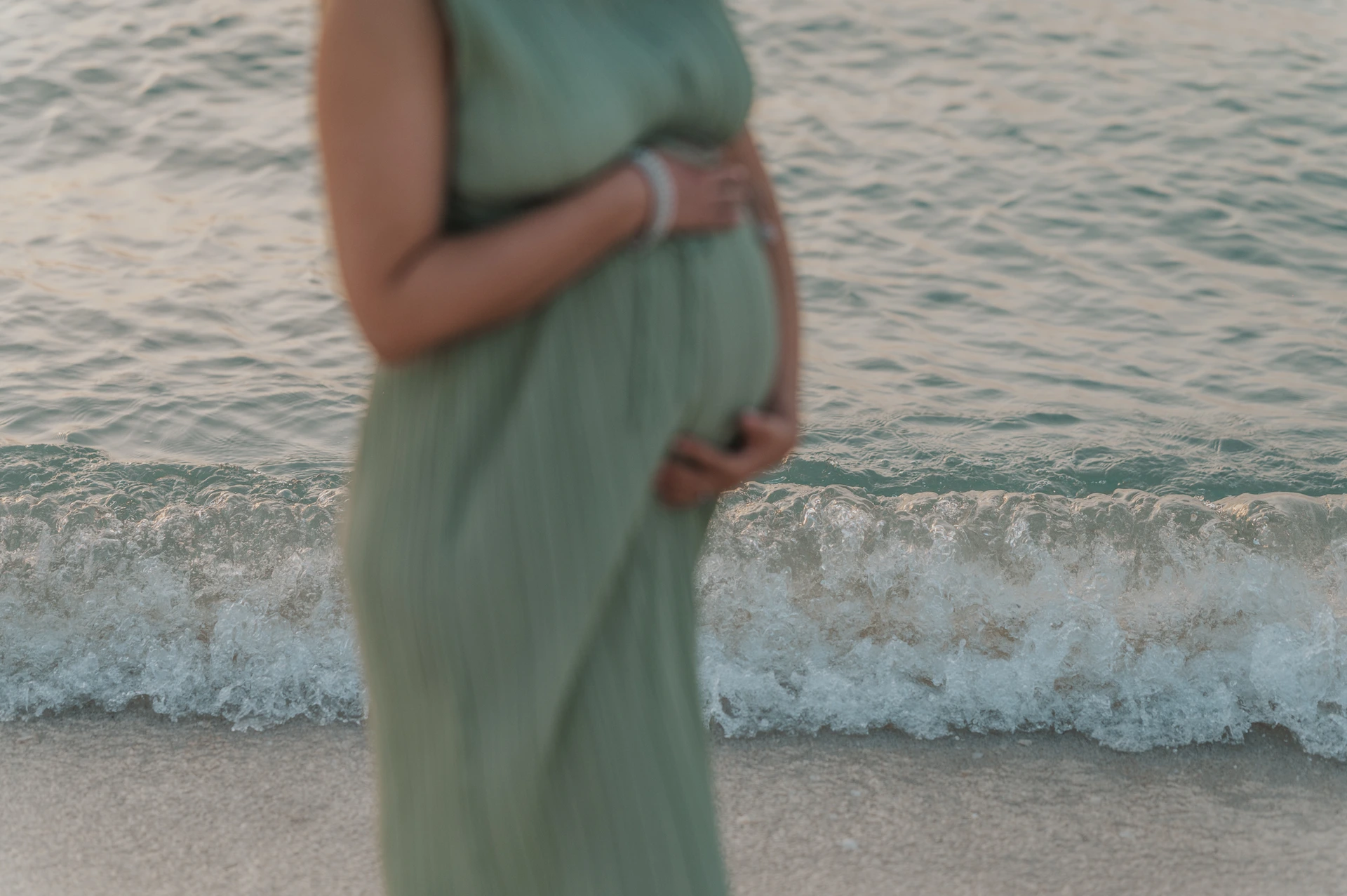 A pregnant woman in a sage-green dress holds her bump in soft focus beside a breaking wave during a Dubai beach maternity photo.
