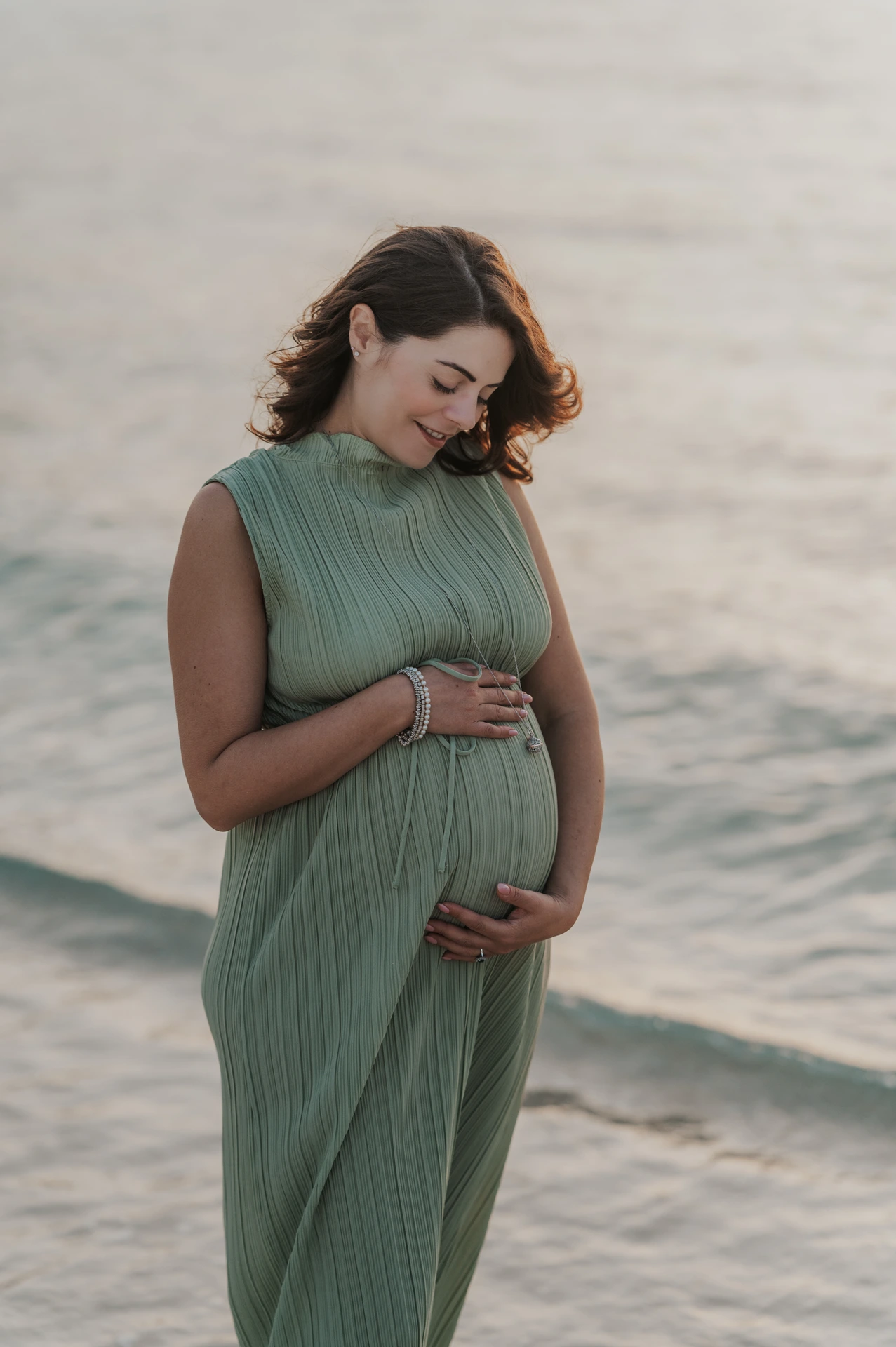 A pregnant woman in a sage-green dress looks down while cradling her bump beside the sea during a Dubai beach maternity portrait.