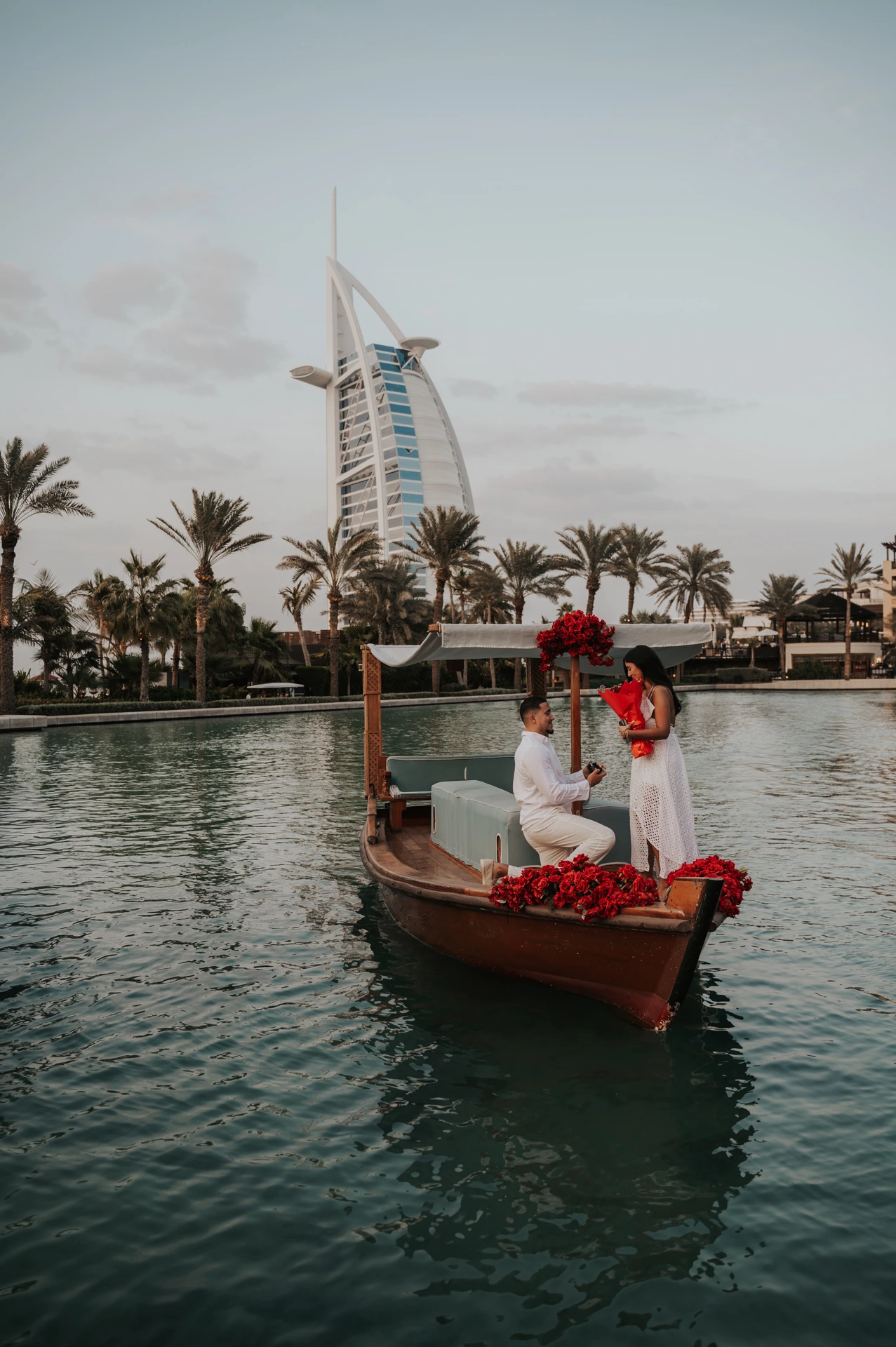 A man kneels with an open ring box on a rose-decorated abra with Burj Al Arab behind their Dubai proposal.
