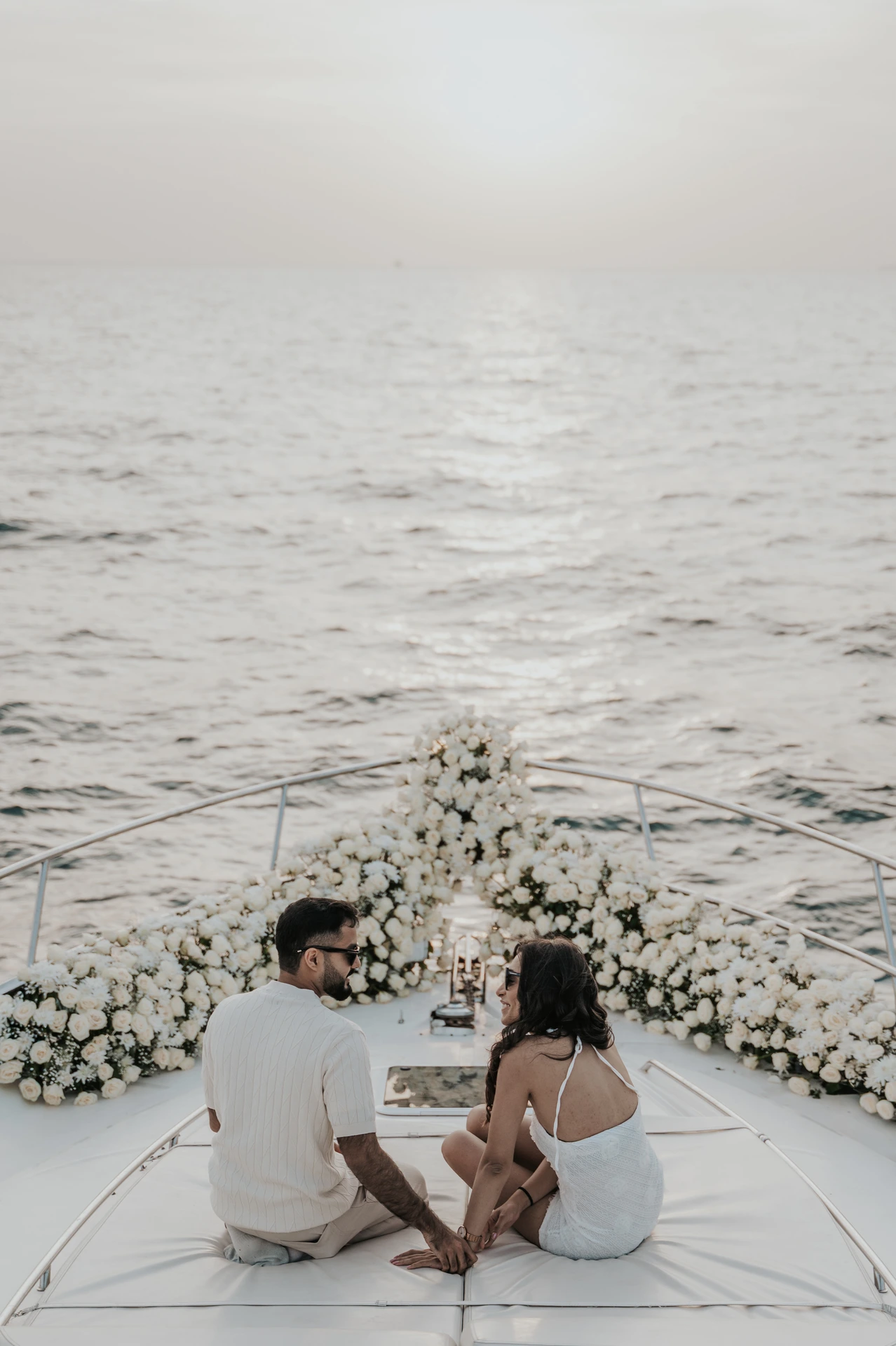 A couple sits hand in hand on a yacht bow surrounded by white flowers after their Dubai proposal at sunset.
