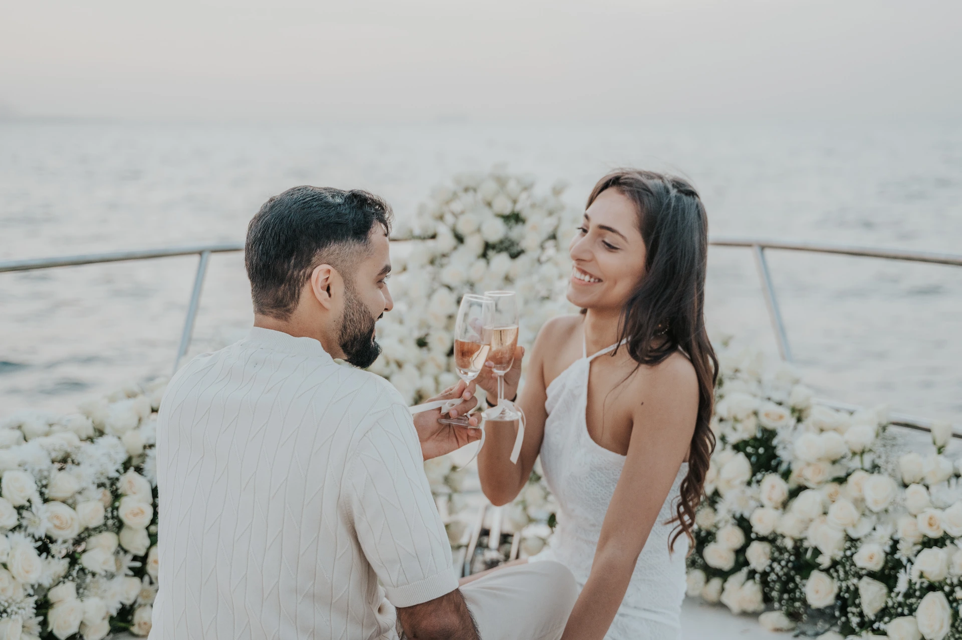 Dubai Yacht Proposal Champagne Toast A couple raises champagne glasses on a yacht bow surrounded by white flowers after their Dubai proposal.