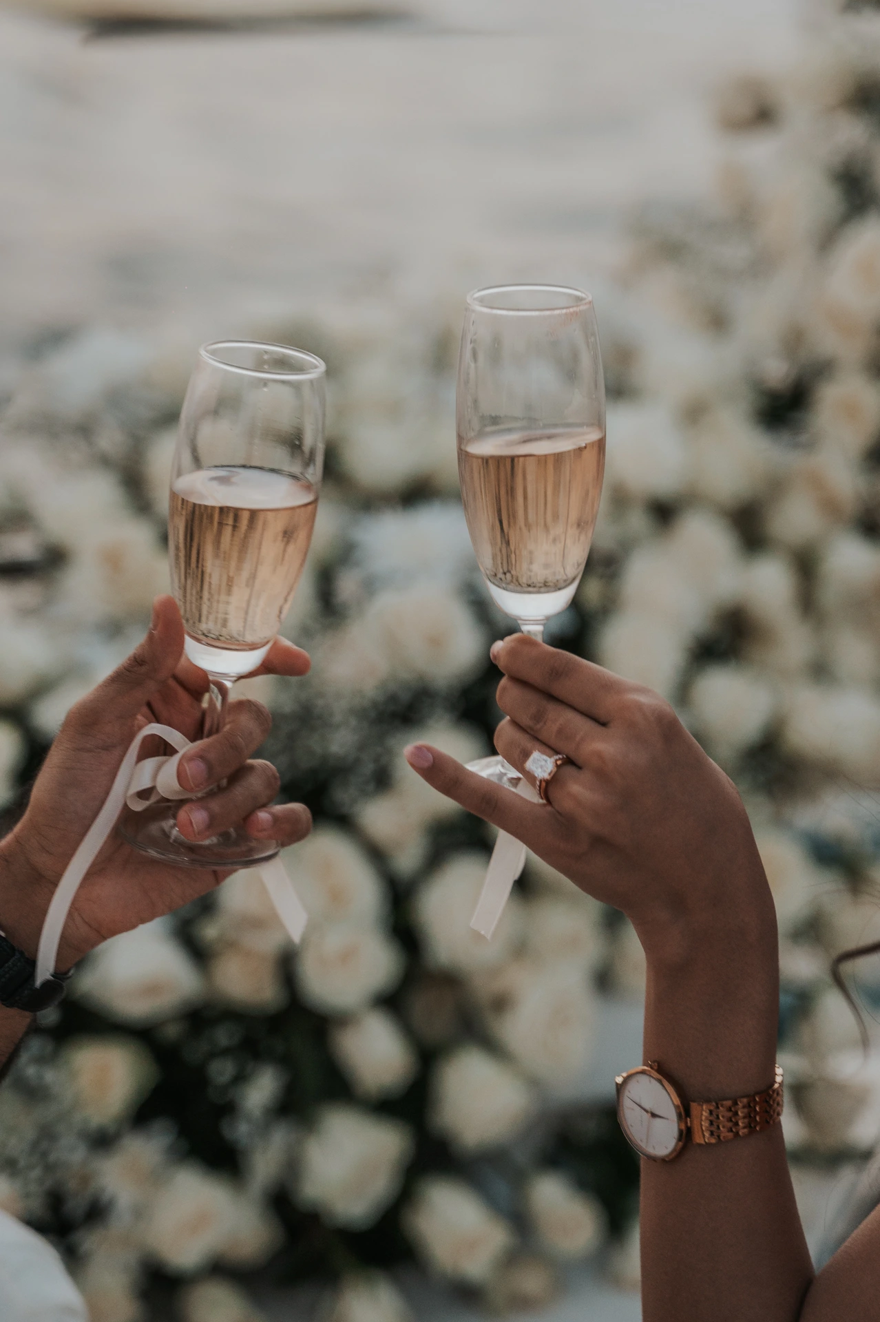 A couple raises champagne glasses while an engagement ring remains visible before white flowers at their Dubai proposal celebration.