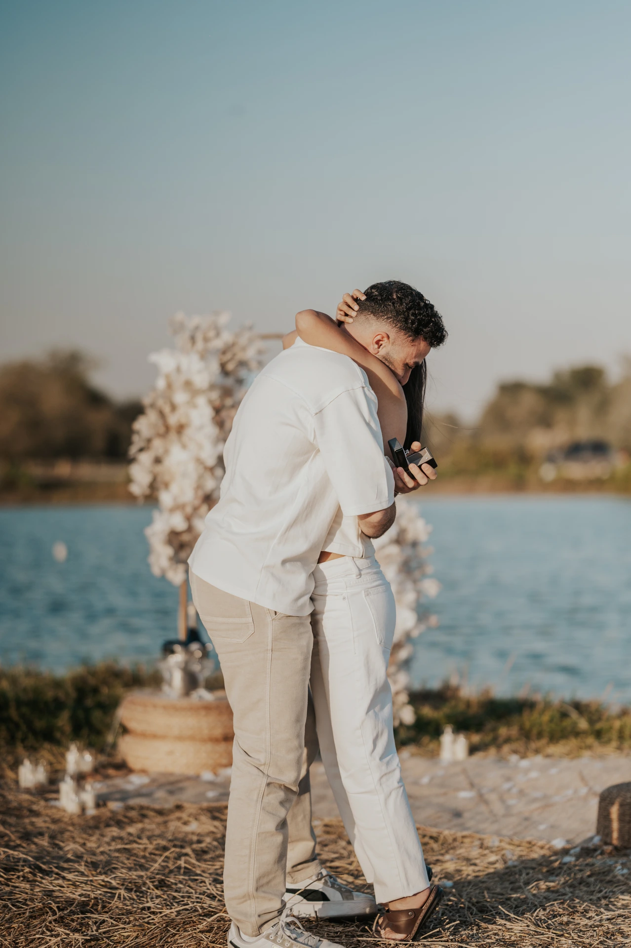 Dubai Lakeside Proposal Hug with Ring Box A couple hugs beside a lakeside floral setup while the man holds an open ring box during a Dubai proposal.