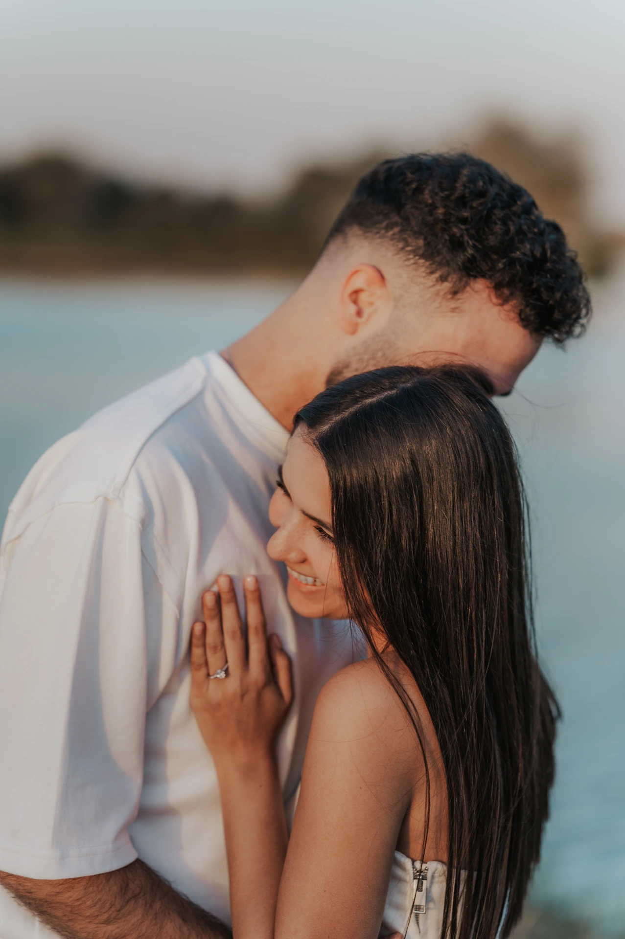 A couple embraces beside the water while the woman displays her ring during a Dubai proposal photography portrait.