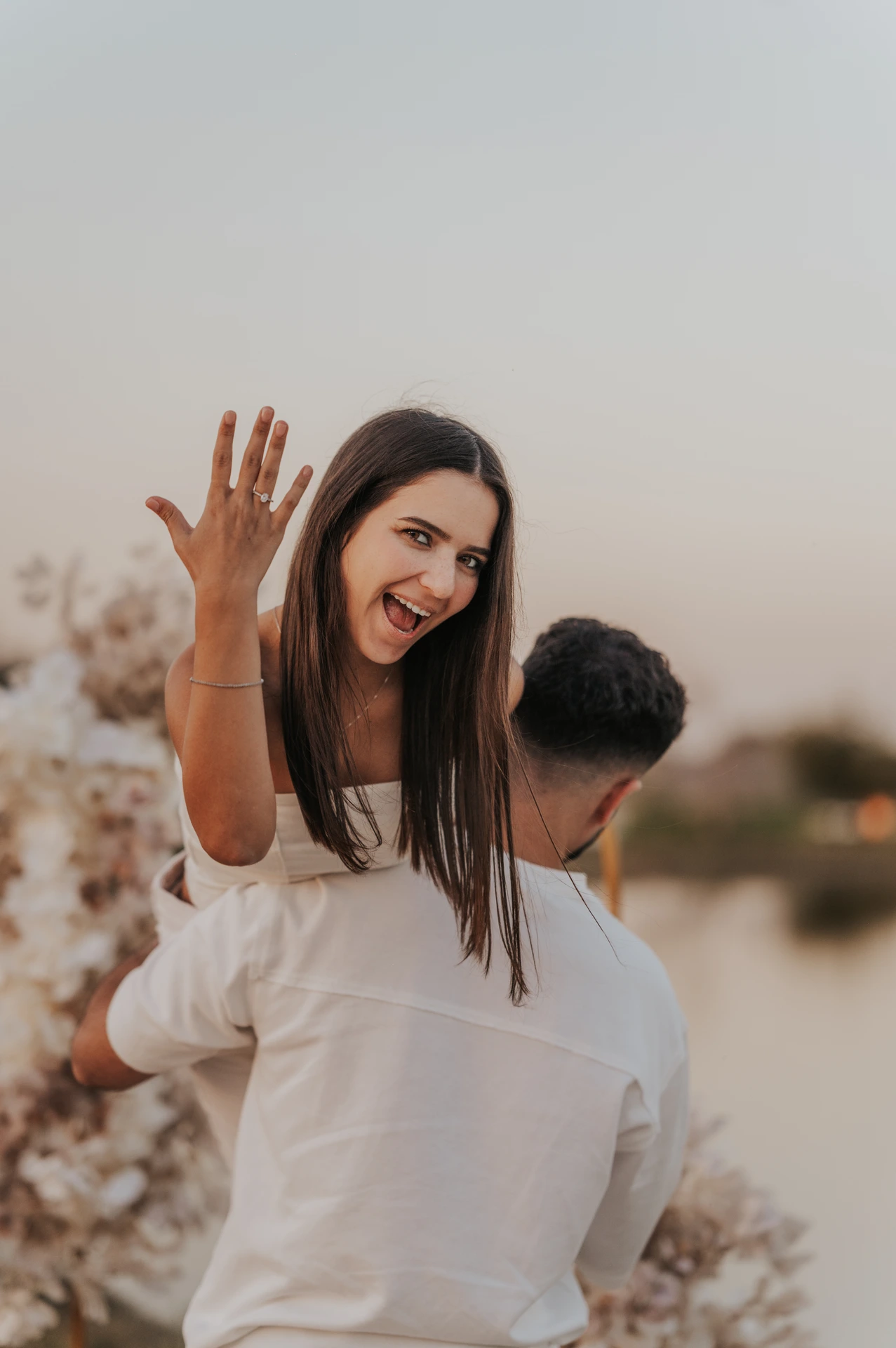 A woman displays her engagement ring while her partner lifts her beside a lakeside floral setup after a Dubai proposal.