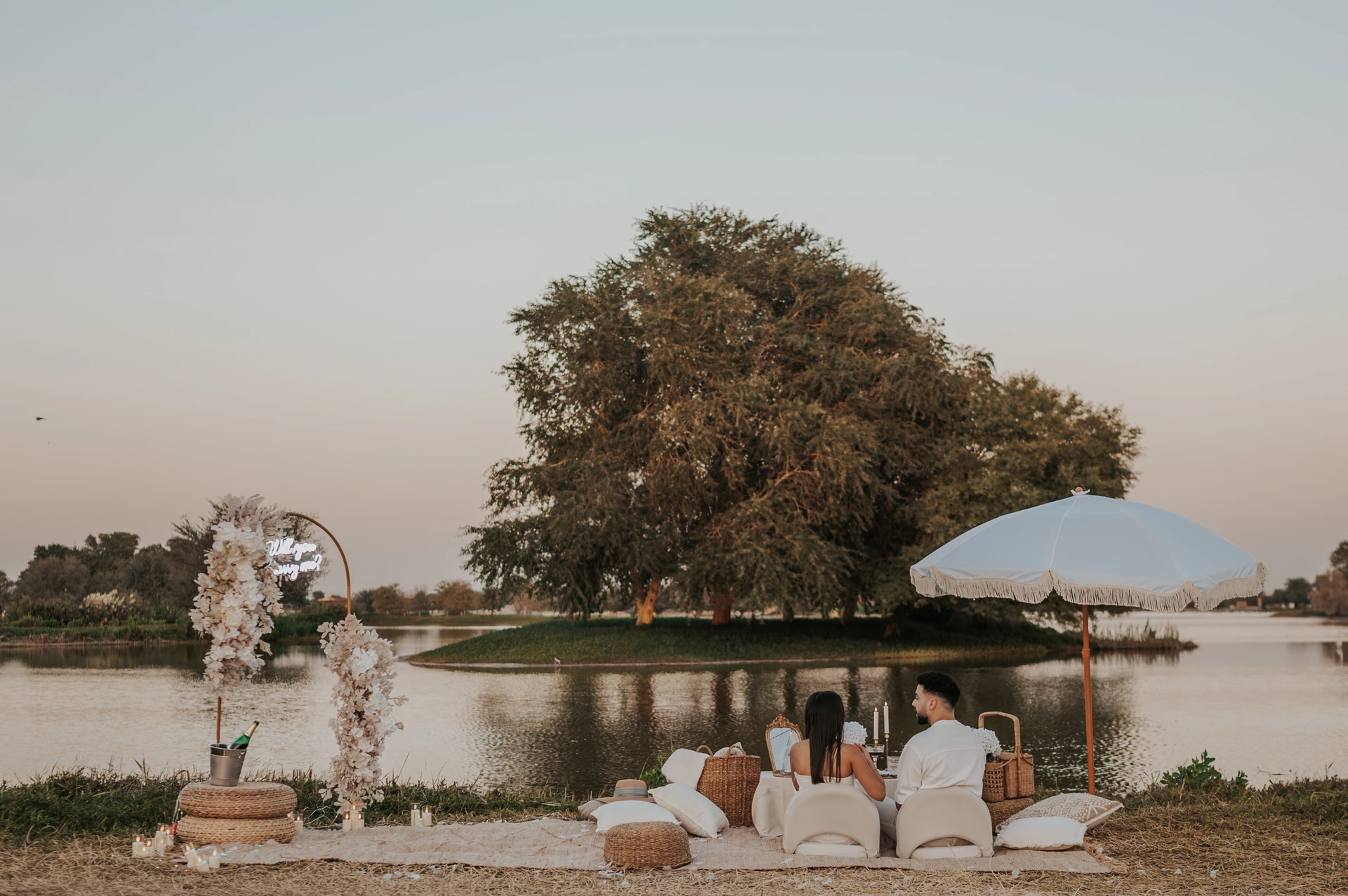 A couple sits at a styled lakeside picnic with a floral arch and parasol during a Dubai proposal photography session.