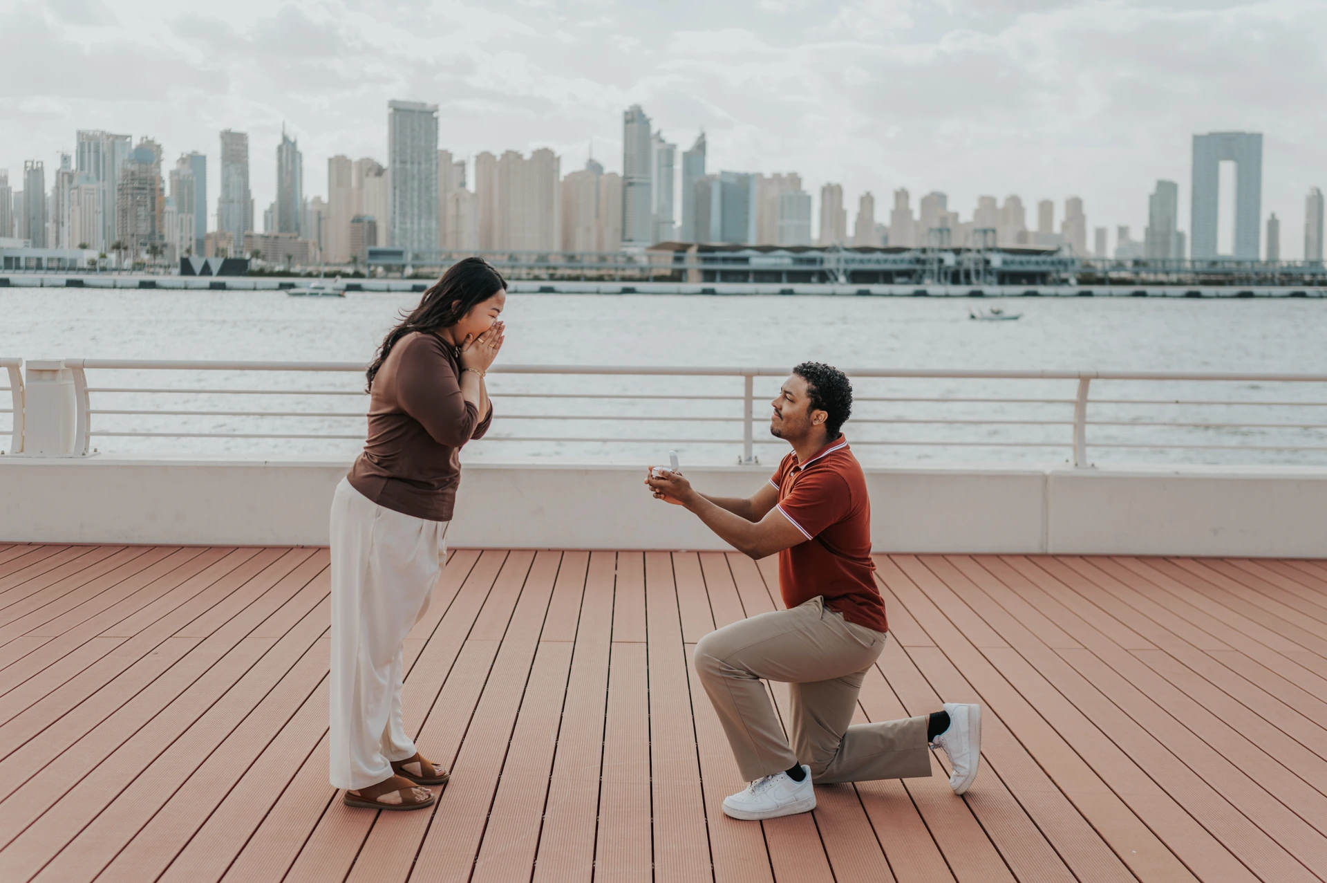 A man kneels with an open ring box on a yacht deck as his partner reacts during a Dubai Marina proposal.