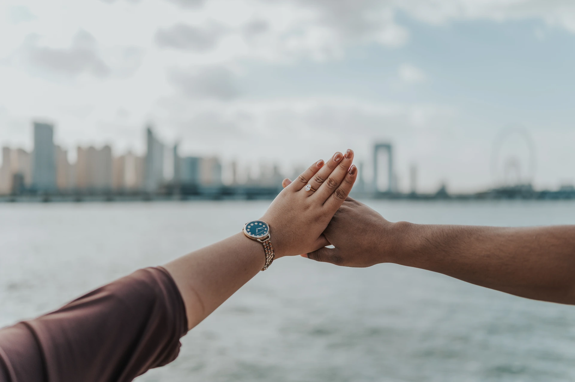 Ain Dubai Proposal Ring Portrait A couple touches hands to display an engagement ring above the water, with Ain Dubai blurred behind their Dubai proposal portrait.