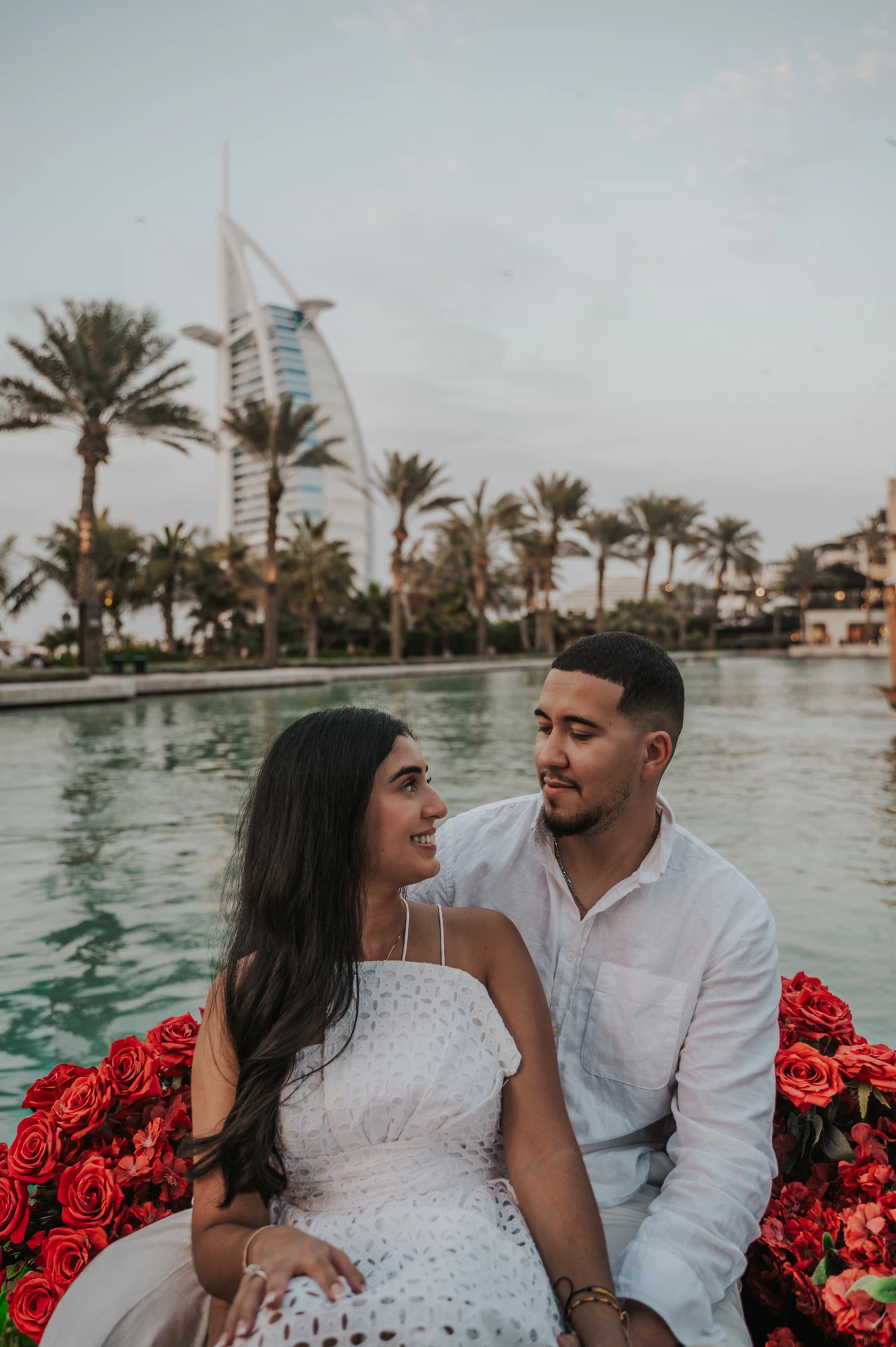 A couple sits together on a rose-filled abra with Burj Al Arab behind their Dubai proposal photographer portrait.