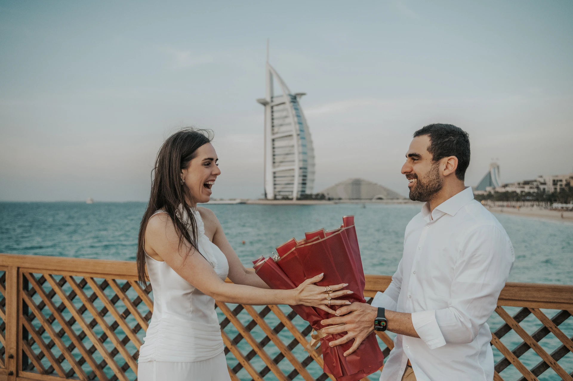 A smiling couple holds a red bouquet by the water with Burj Al Arab behind them after a Dubai proposal.