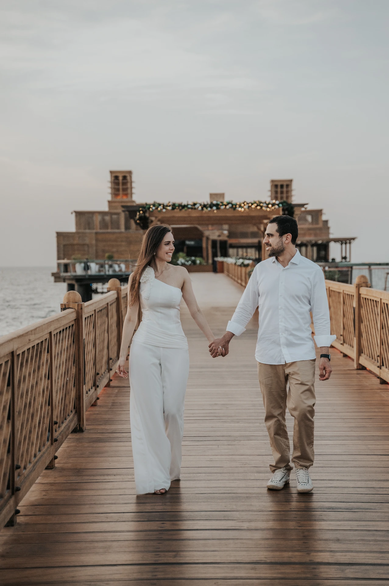 A couple walks hand in hand along a wooden resort pier during their Dubai proposal photographer portrait session.