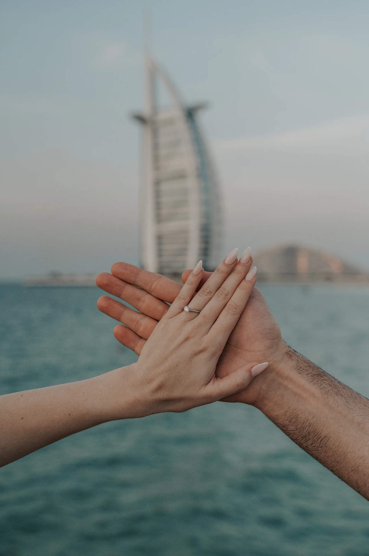A couple holds their hands together to show an engagement ring with Burj Al Arab behind their Dubai proposal portrait.
