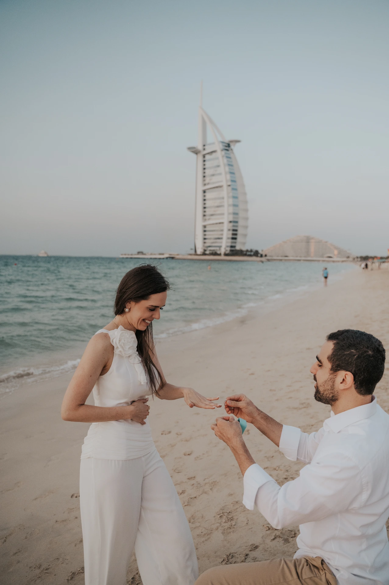 A man kneels and holds an open ring box on the beach with Burj Al Arab behind their Dubai proposal.