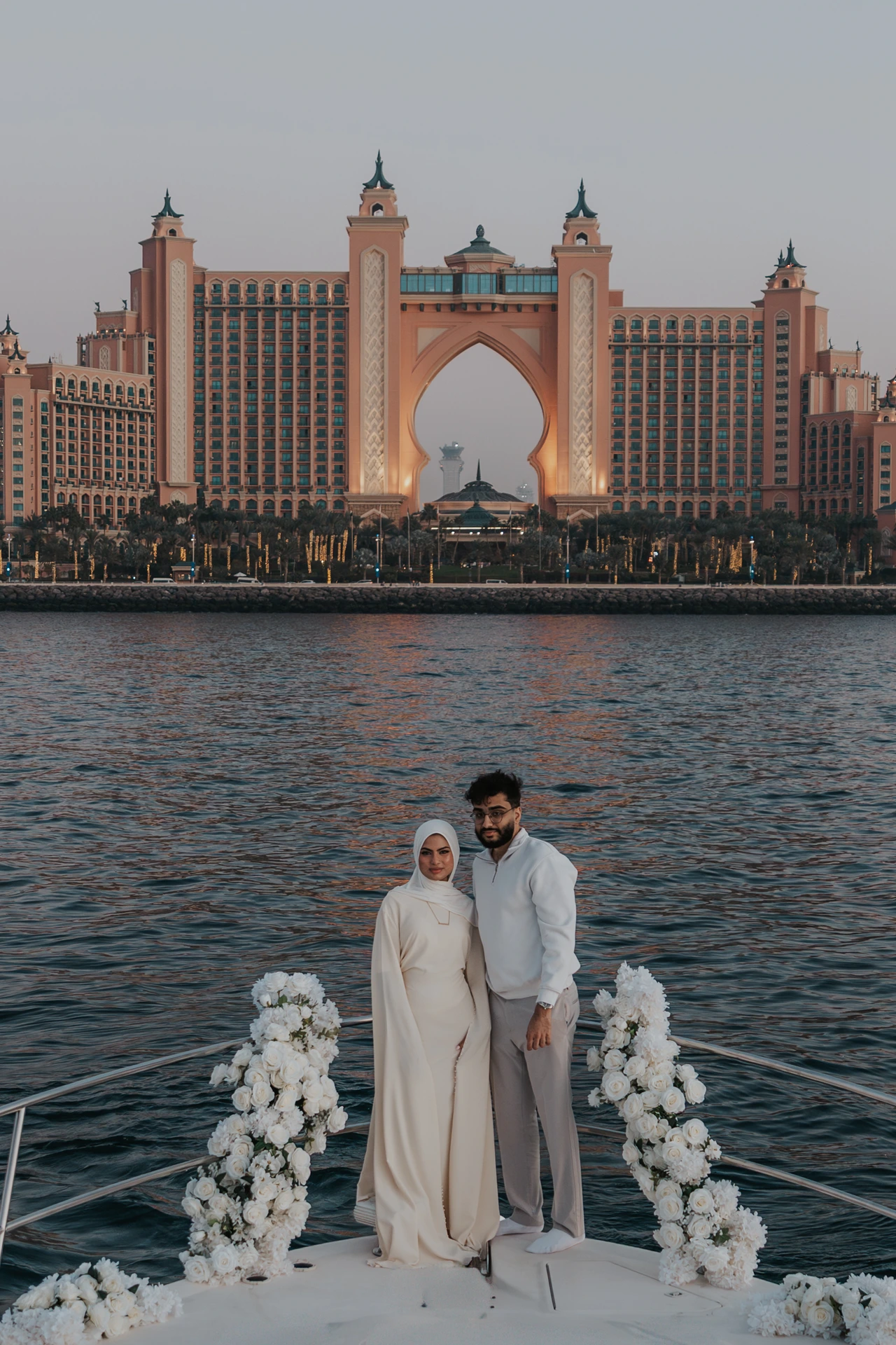 A couple poses between white floral arrangements on a yacht bow with Atlantis The Palm behind their Dubai proposal portrait.