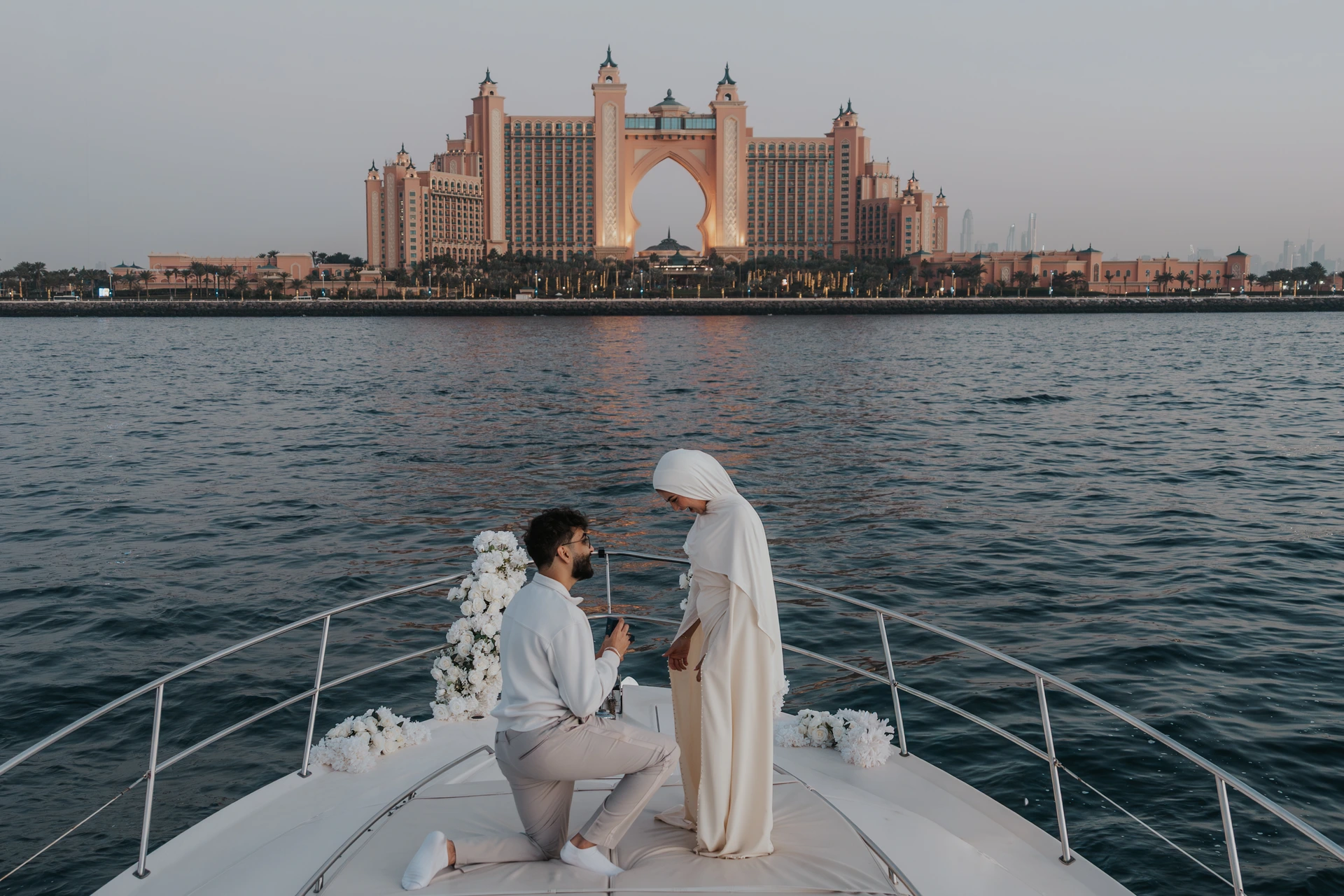 A man kneels on a flower-decorated yacht bow with Atlantis The Palm behind his partner during a Dubai proposal.