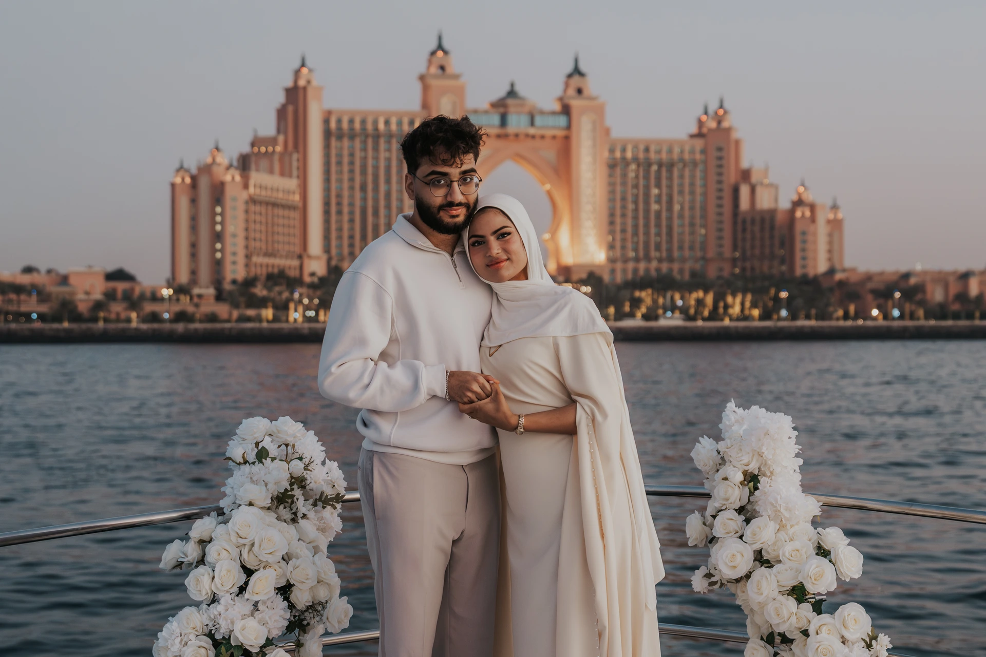 A couple poses between white flowers on a yacht with Atlantis The Palm behind them after their Dubai proposal.