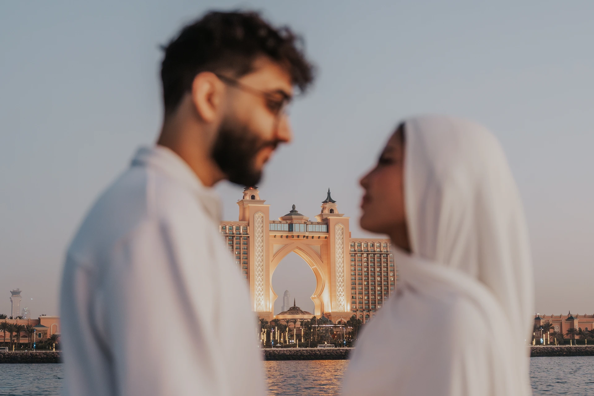 A couple in white stands close together with Atlantis The Palm behind them during a Dubai proposal photographer session.