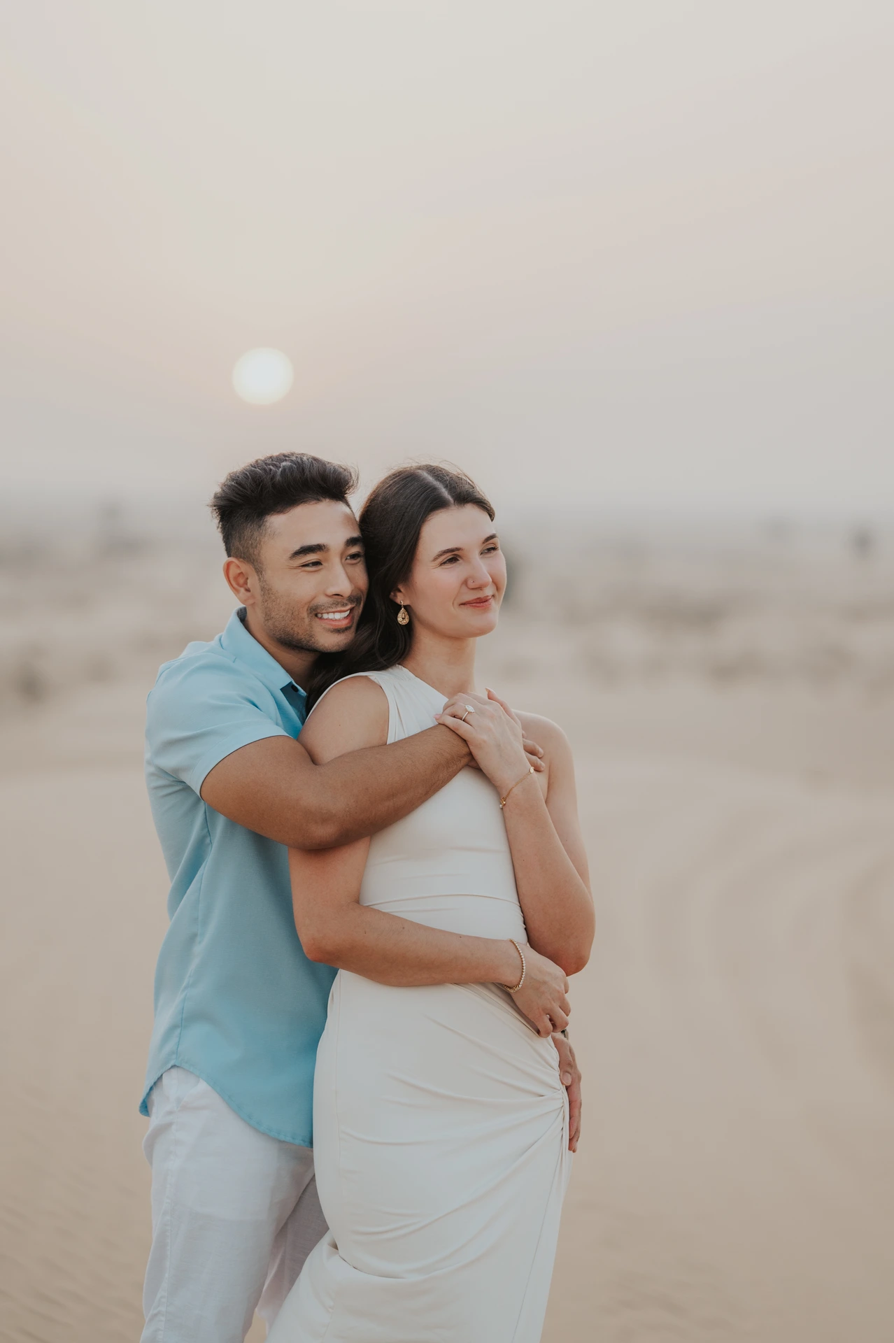 A man embraces his partner from behind on desert dunes at sunset during their Dubai proposal photographer portrait.