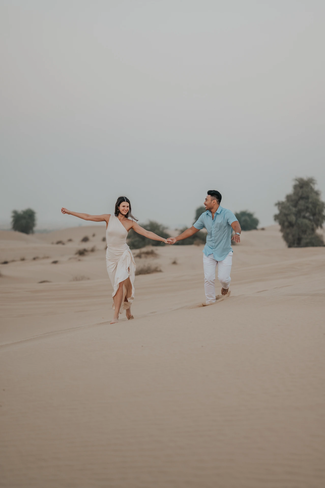 A couple runs hand in hand across desert dunes during a playful portrait after their Dubai proposal photoshoot.
