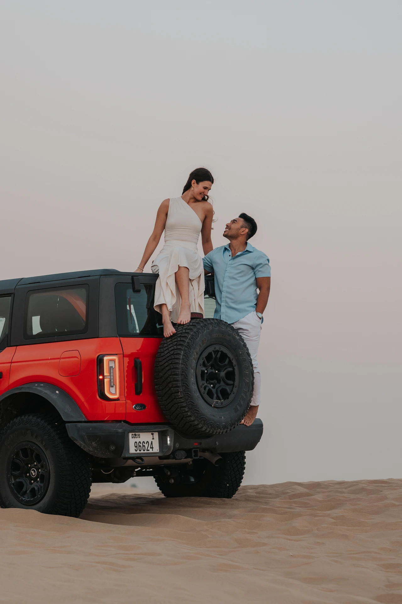 A couple poses together on the back of a red Ford Bronco among the dunes during a Dubai proposal photoshoot.