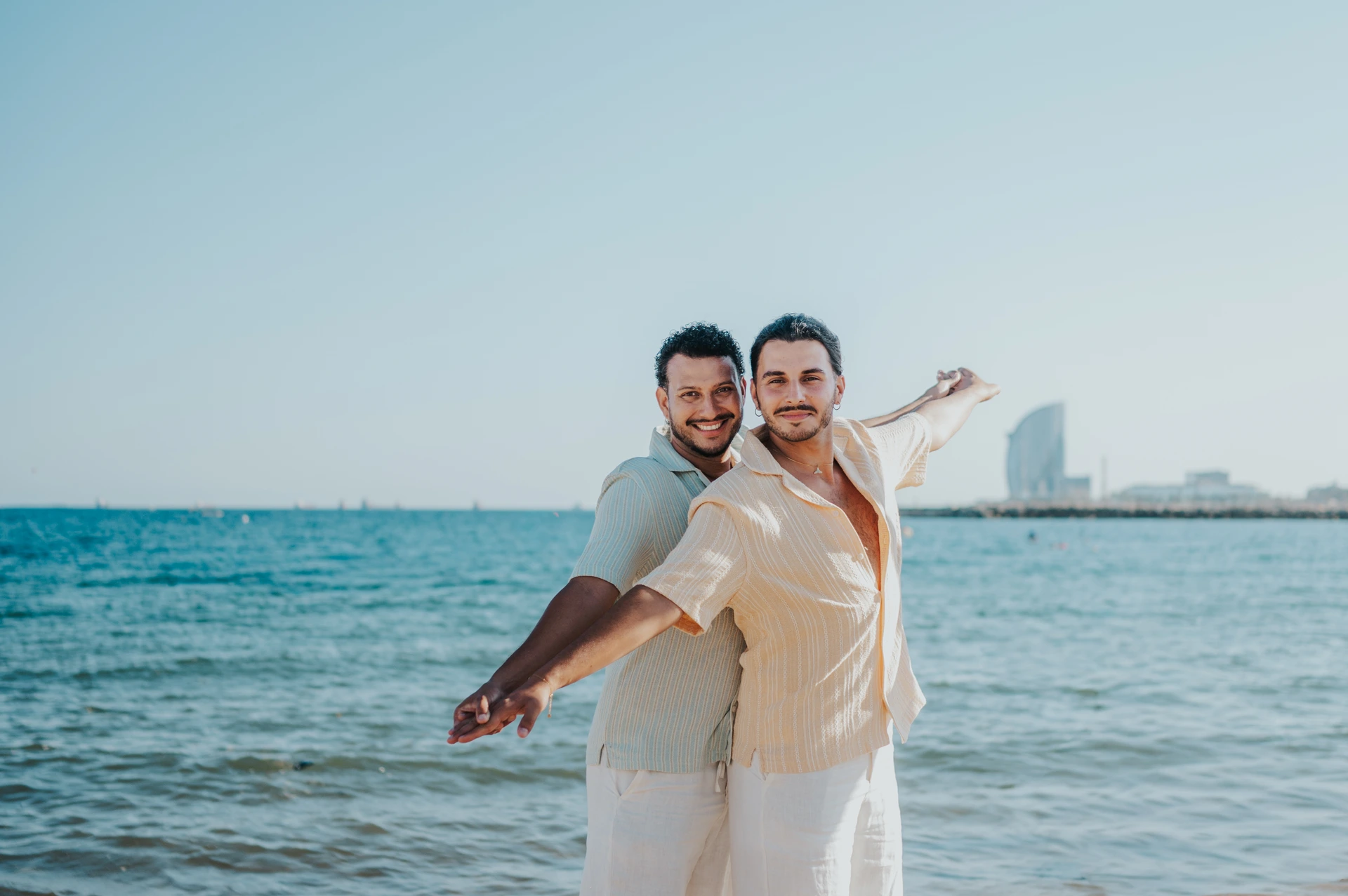 Twee mannen poseren samen met uitgestrekte armen op het Barceloneta-strand terwijl W Barcelona in de verte zichtbaar is tijdens een fotoshoot van 90 minuten in Barcelona.