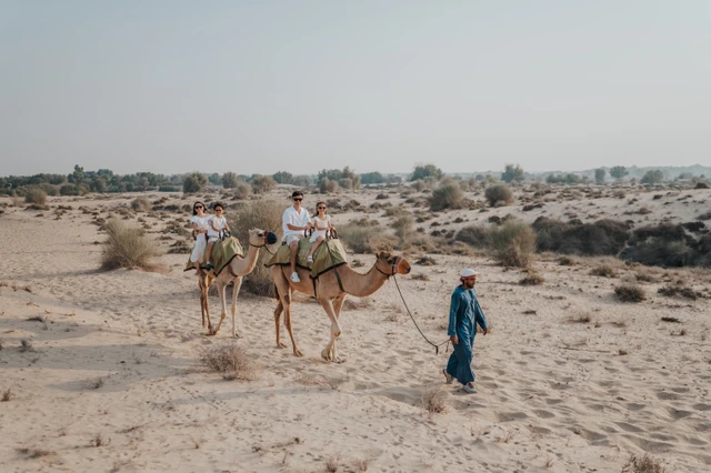 A family walks beside camels on Dubai desert sand during a vacation family photoshoot, capturing an adventurous travel moment with open dunes and soft light.