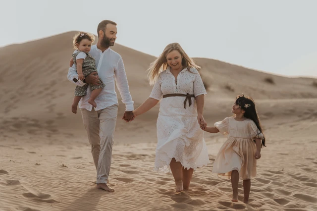A family walks together across Dubai desert dunes during a vacation family photoshoot, creating a natural movement portrait with warm neutral tones.