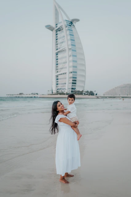 A mother holds her toddler near Dubai beach during a vacation family photoshoot, with Burj Al Arab and calm sea creating a soft coastal backdrop.