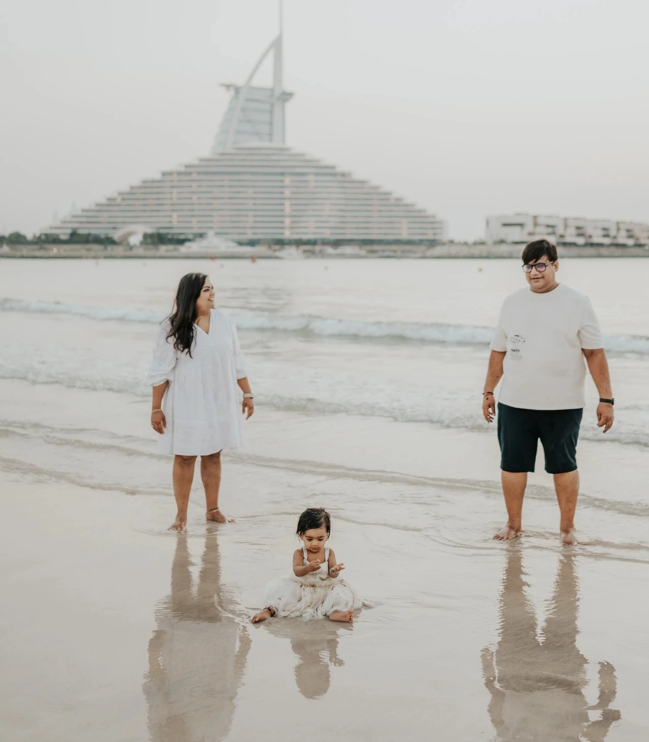 Parents stand with their child at Dubai shoreline during a vacation family photoshoot, with Burj Al Arab visible behind them in a relaxed beach frame.