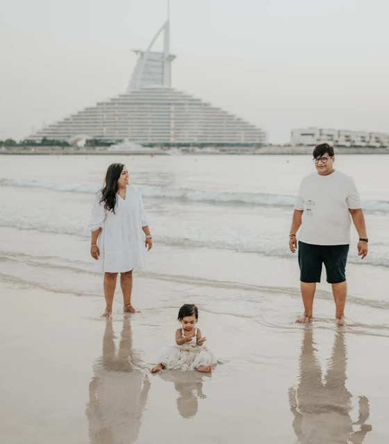 Parents stand with their child at Dubai shoreline during a vacation family photoshoot, with Burj Al Arab visible behind them in a relaxed beach frame.