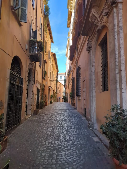 Via della Reginella in Rome A quiet cobbled lane runs between tall ochre and terracotta facades with shutters and small plants in Rome.