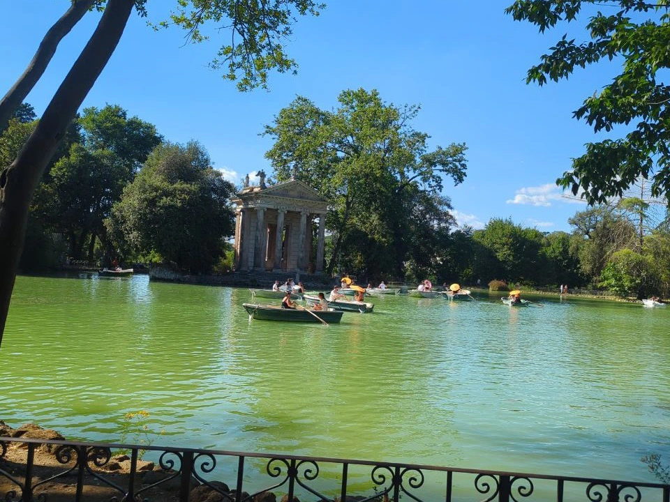 Rowboats cross the green Villa Borghese lake beneath mature trees, showing a calm park setting for a Rome maternity photoshoot.