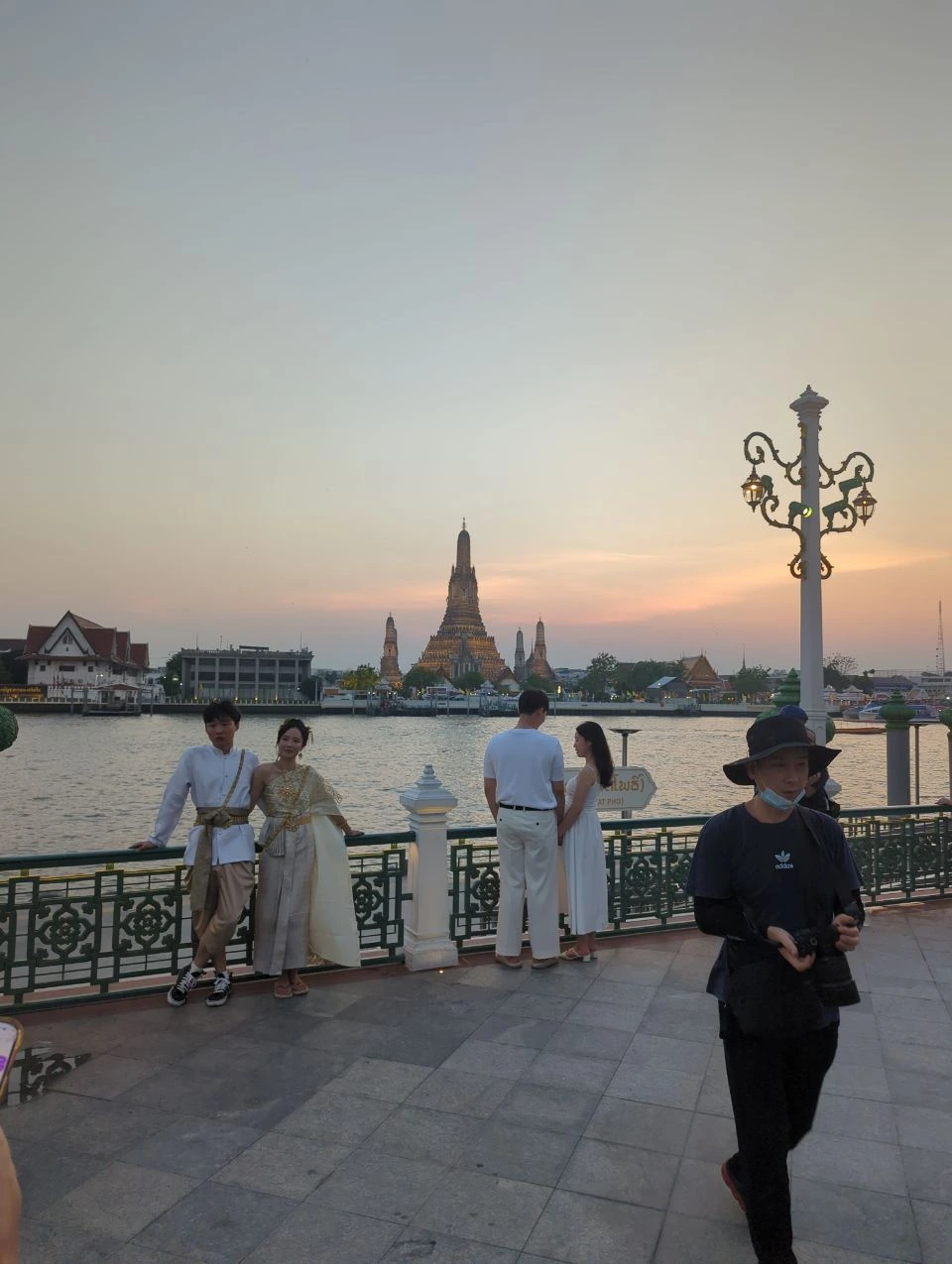 Wat Pho vs Wat Arun Photos - Evening Light Visitors gather beside Bangkok’s river as illuminated Wat Arun stands across the water beneath a warm evening sky.