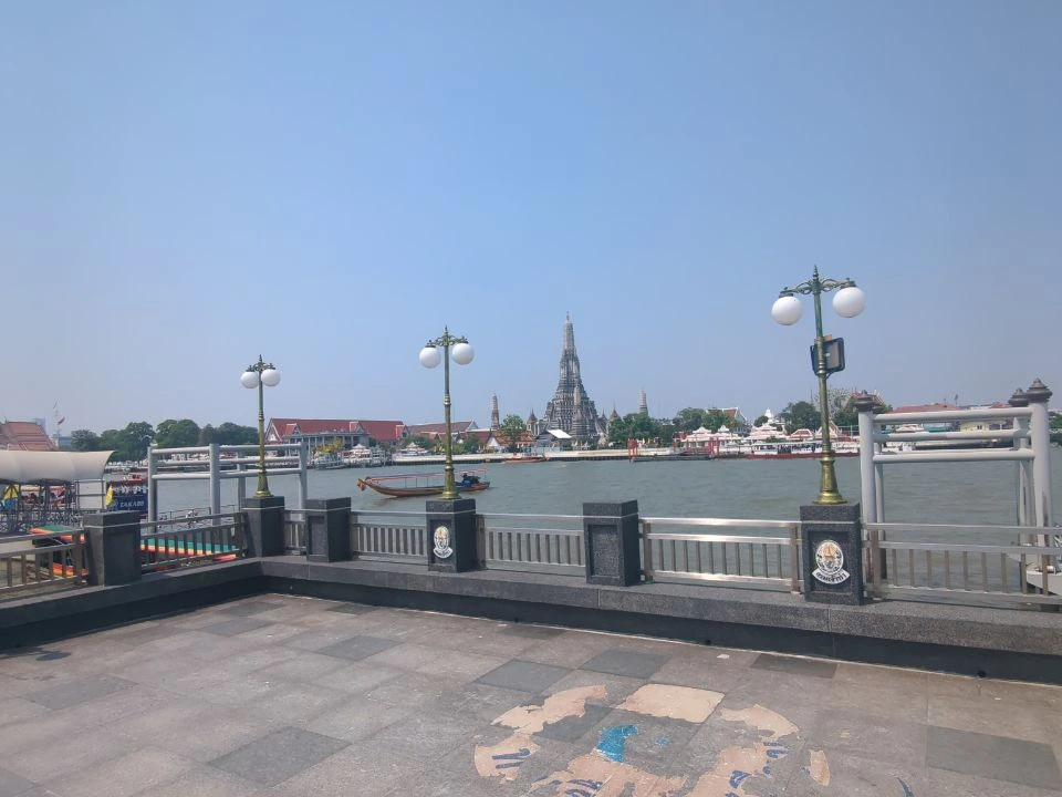 Wat Arun rises across the Chao Phraya River beyond an open Bangkok promenade, with a small boat passing in clear daylight.