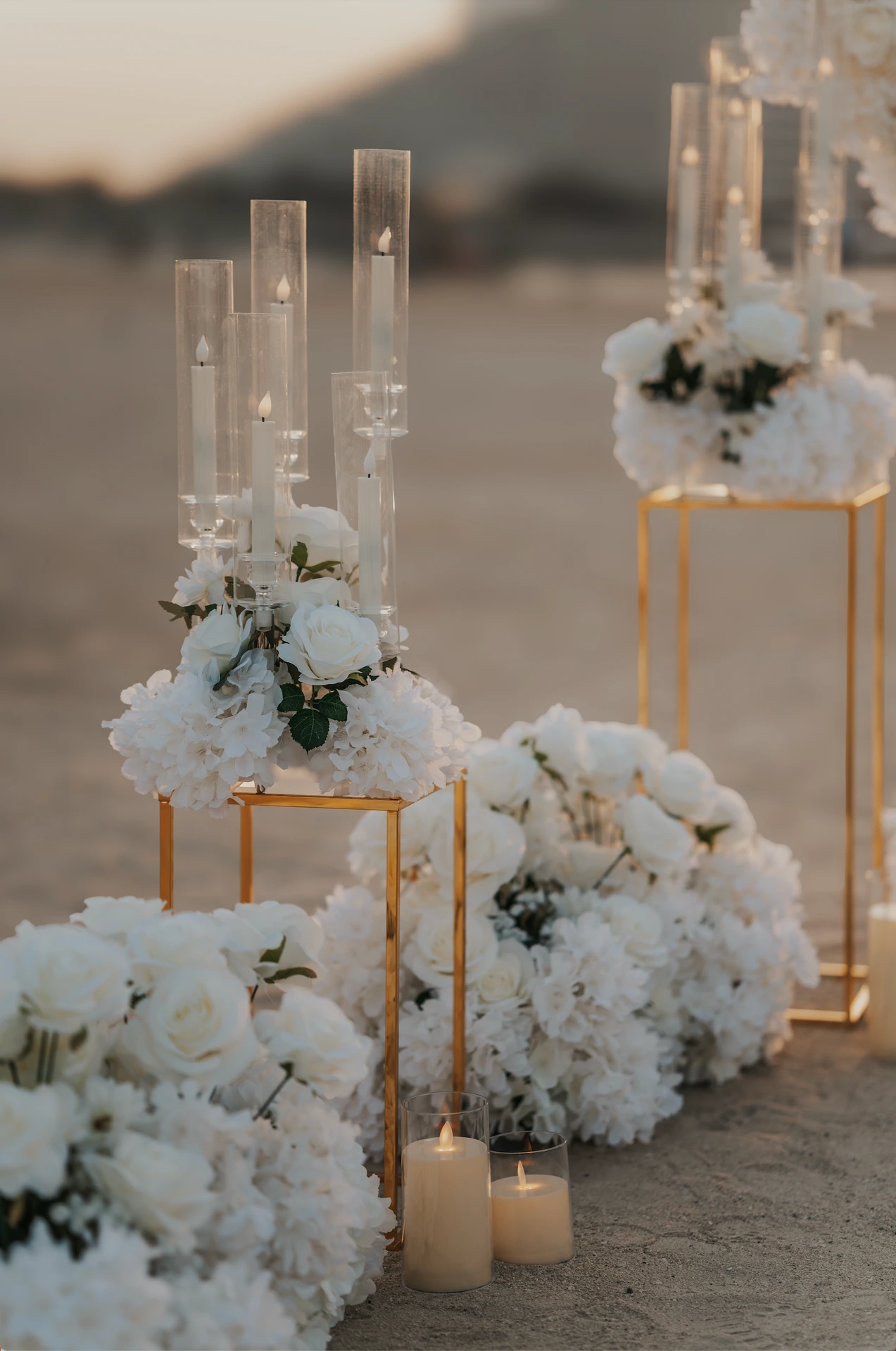 Close-up of white floral arrangements and glass candle holders on Dubai beach, styled as part of a heart proposal setup at golden hour.