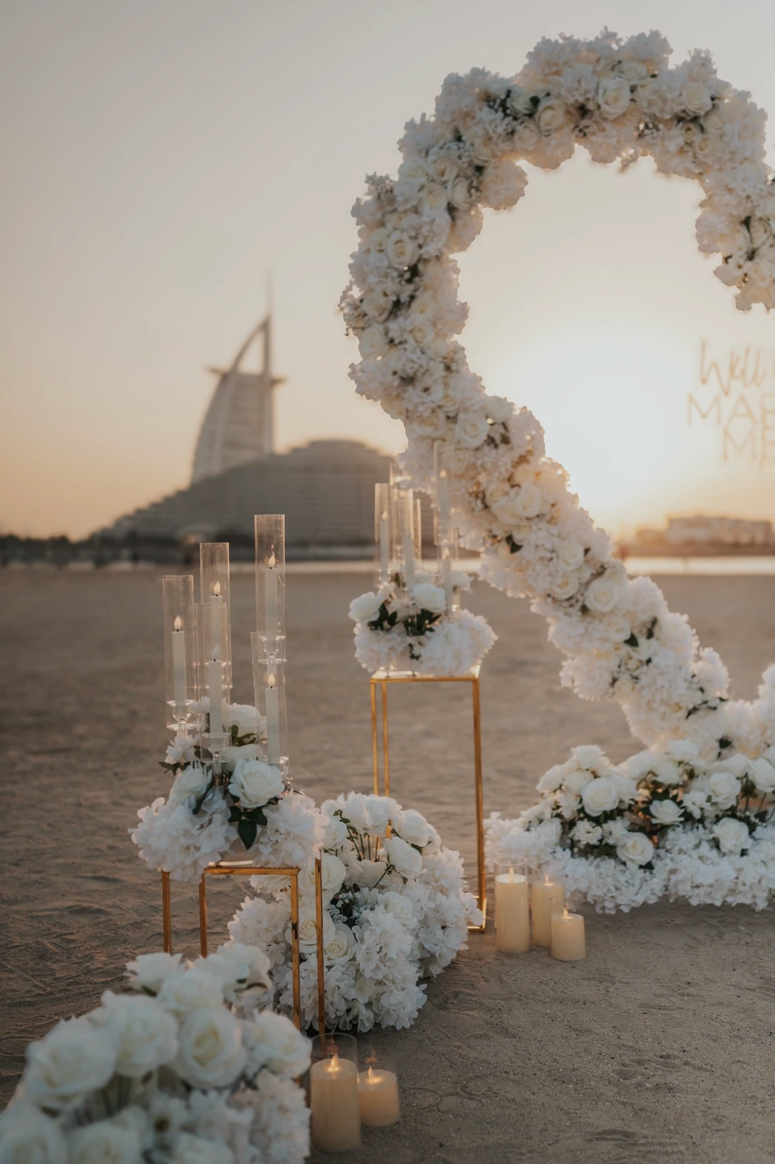 Heart Proposal Setup Dubai - white heart arch at sunset A white floral heart arch stands on Dubai beach during sunset, with candlelight decor and Burj Al Arab in the background for a proposal setup.