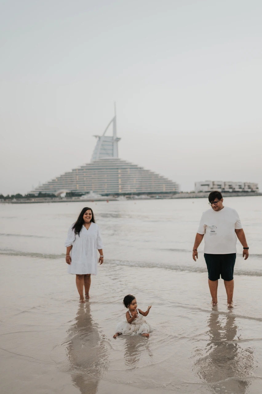 Vacation Family Photoshoot Dubai - Burj beach walk Parents and child walk along Dubai shoreline during a vacation family photoshoot, with Burj Al Arab in the background and gentle sea texture behind.
