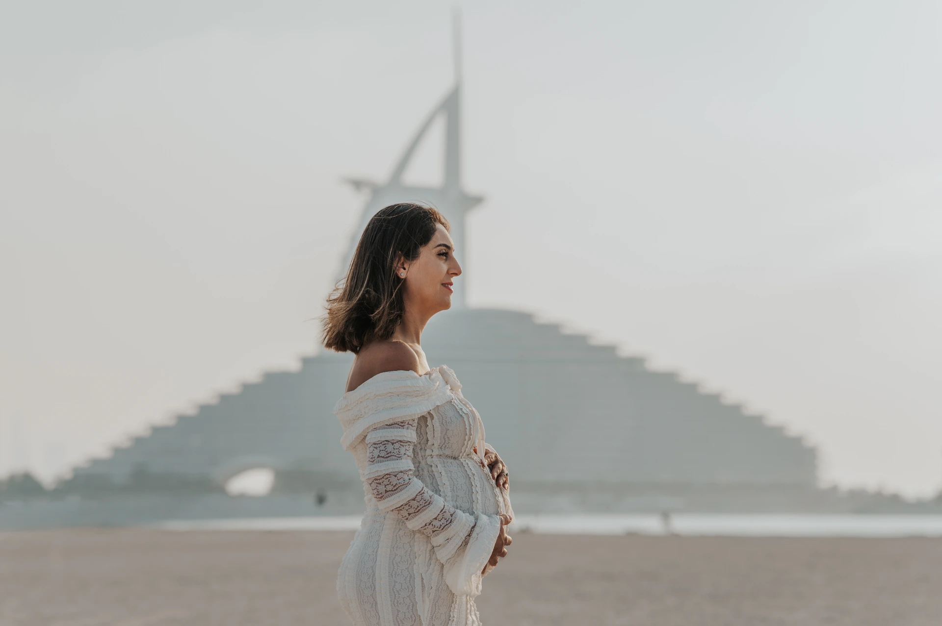 An expecting mother stands in a soft-toned dress on Dubai beach with Burj Al Arab behind her during a maternity photoshoot wardrobe example.